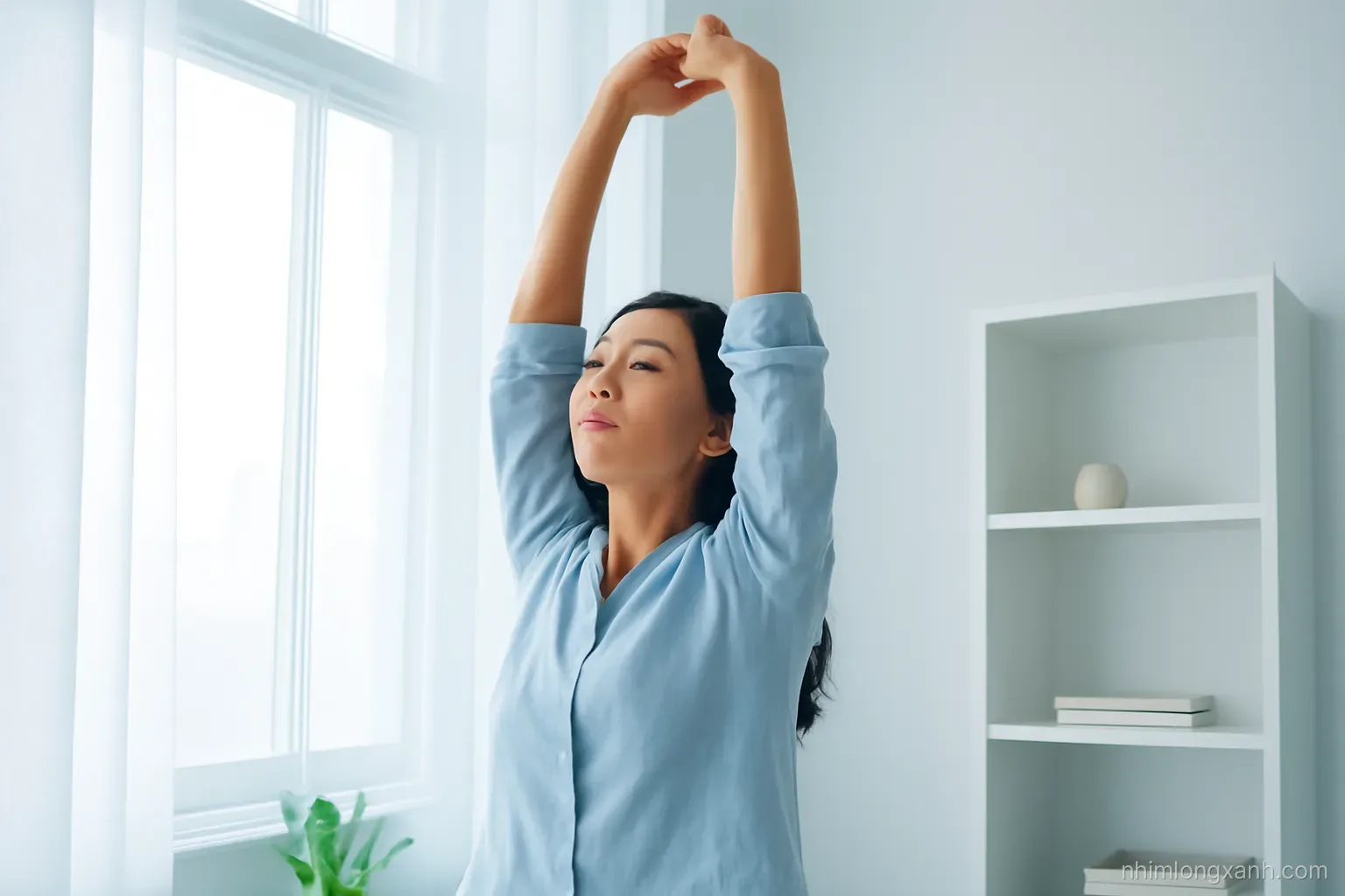 Low angle view of a Vietnamese woman in her thirties stretching her fingers at a bright window in a calm apartment, soft morning haze, pale blue and white tones