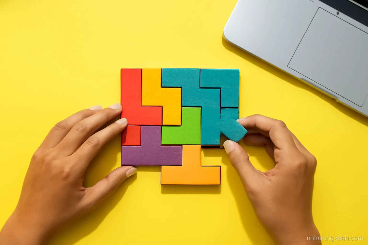 Top-down view of colorful wooden puzzle pieces being fitted together by Vietnamese hands on a bright yellow desk next to a closed laptop, crisp daylight