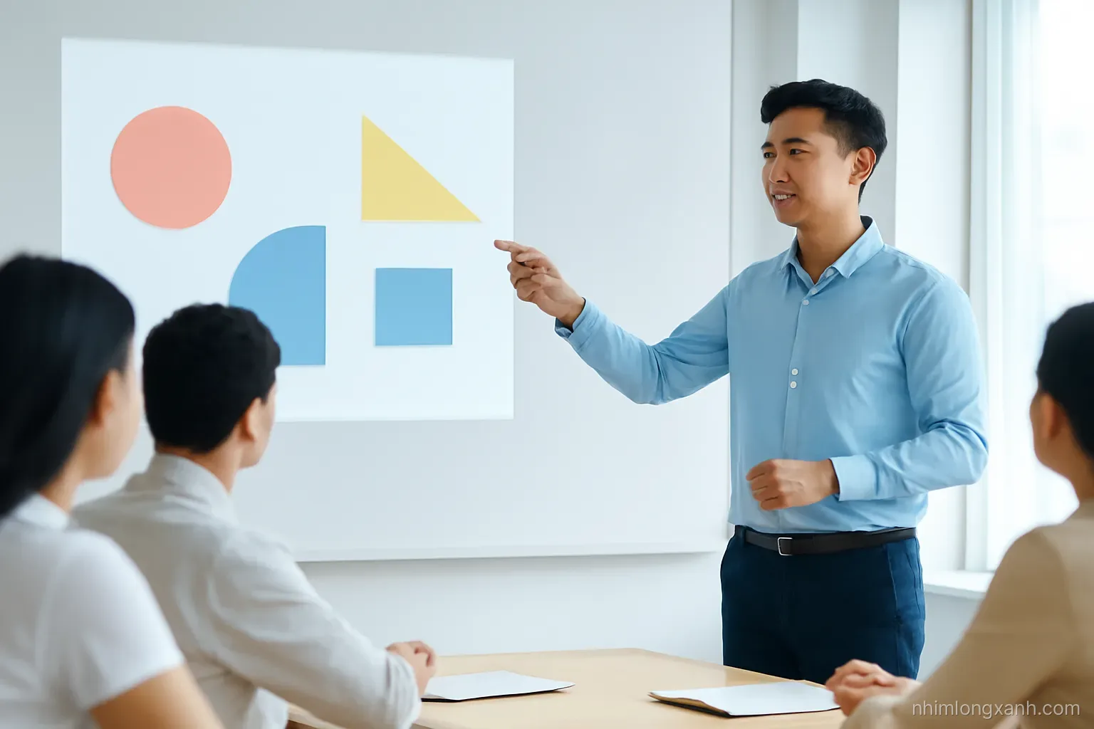 Young Vietnamese man in a light blue shirt presenting beside a projector beam with abstract shapes in a small lecture room, side angle medium shot, three blurred listeners at a table, cool morning light