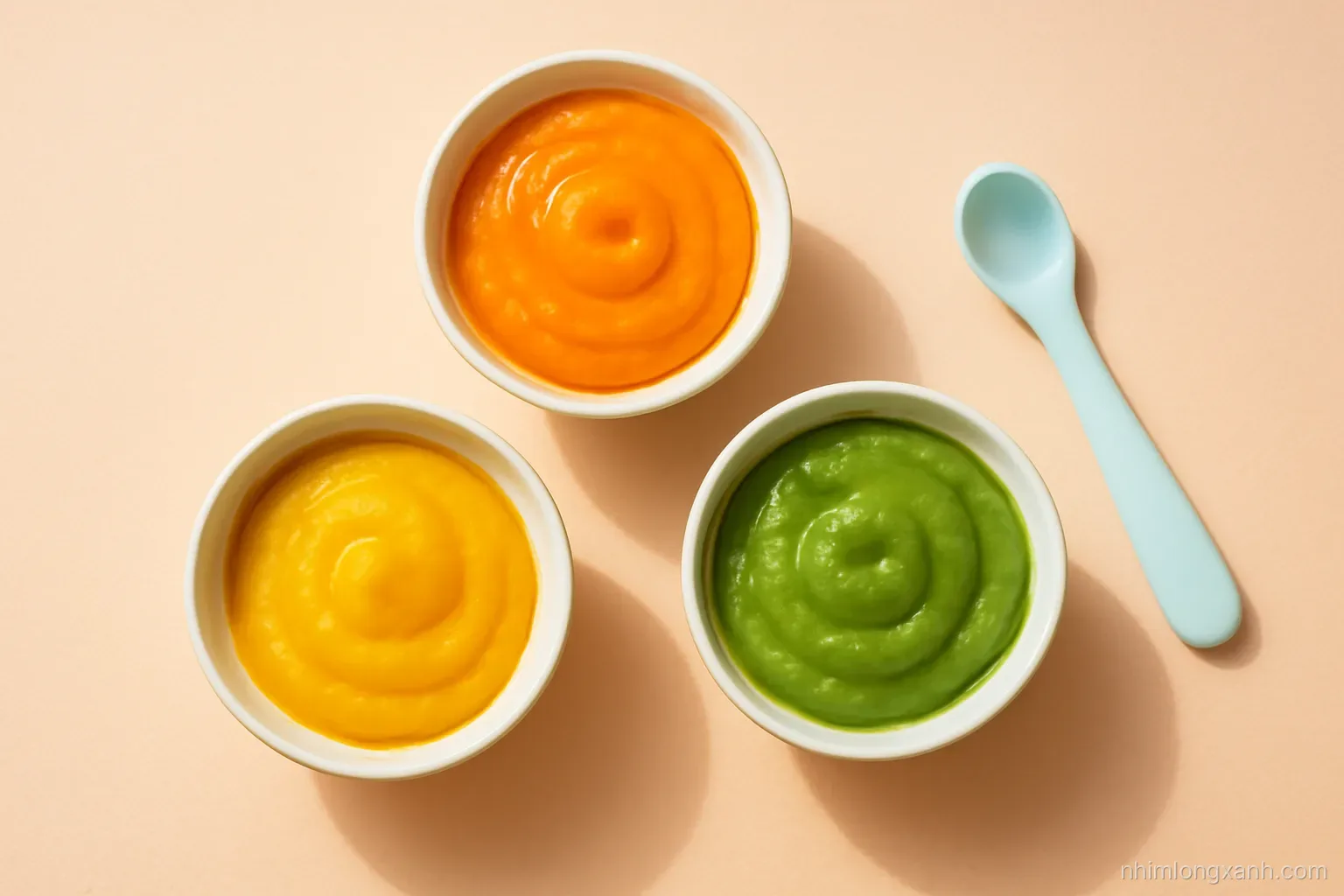 Top-down flat lay of small ceramic bowls with smooth pumpkin, carrot and green pea purees beside a silicone baby spoon on a peach table, bright natural light