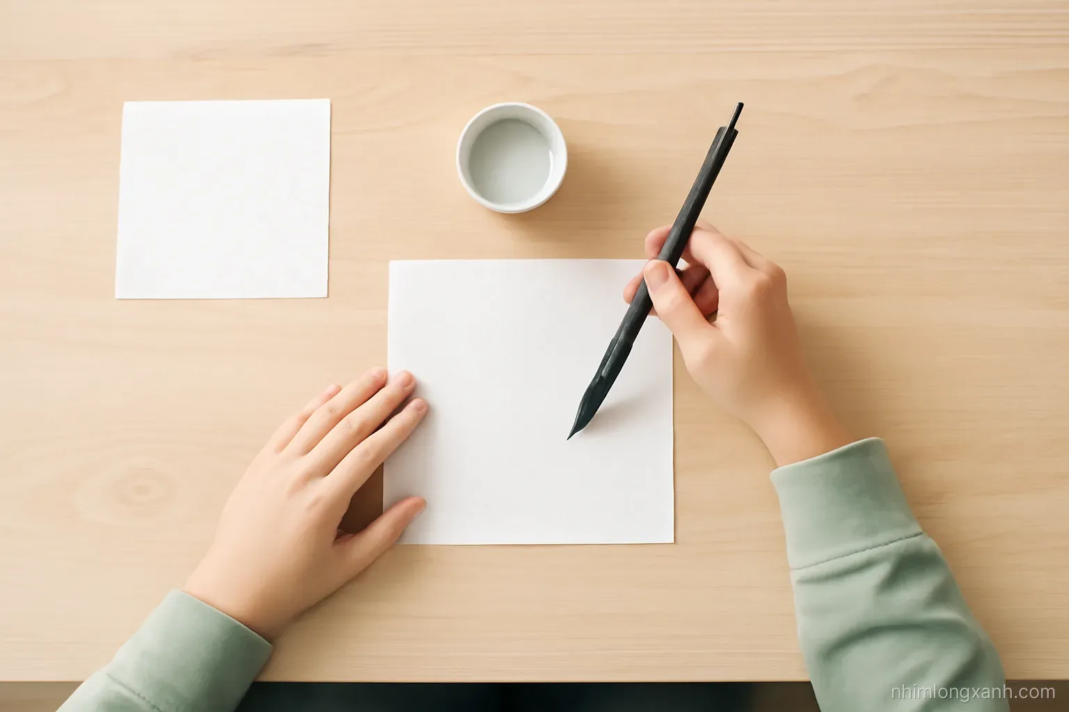 Top-down flat lay of an ink brush, small water dish and blank rice paper squares on a light wooden table, hands of a Vietnamese student, crisp daylight, black and sage green