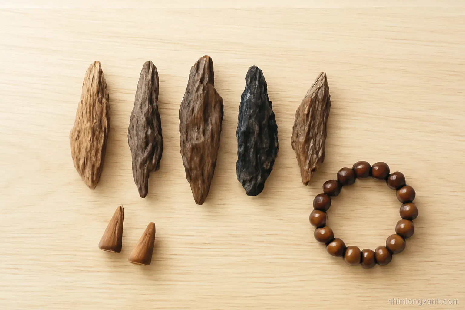 Overhead shot of several agarwood pieces of varying darkness arranged in a row on a light oak table, with incense cones and a bead bracelet, bright airy daylight, brown and cream