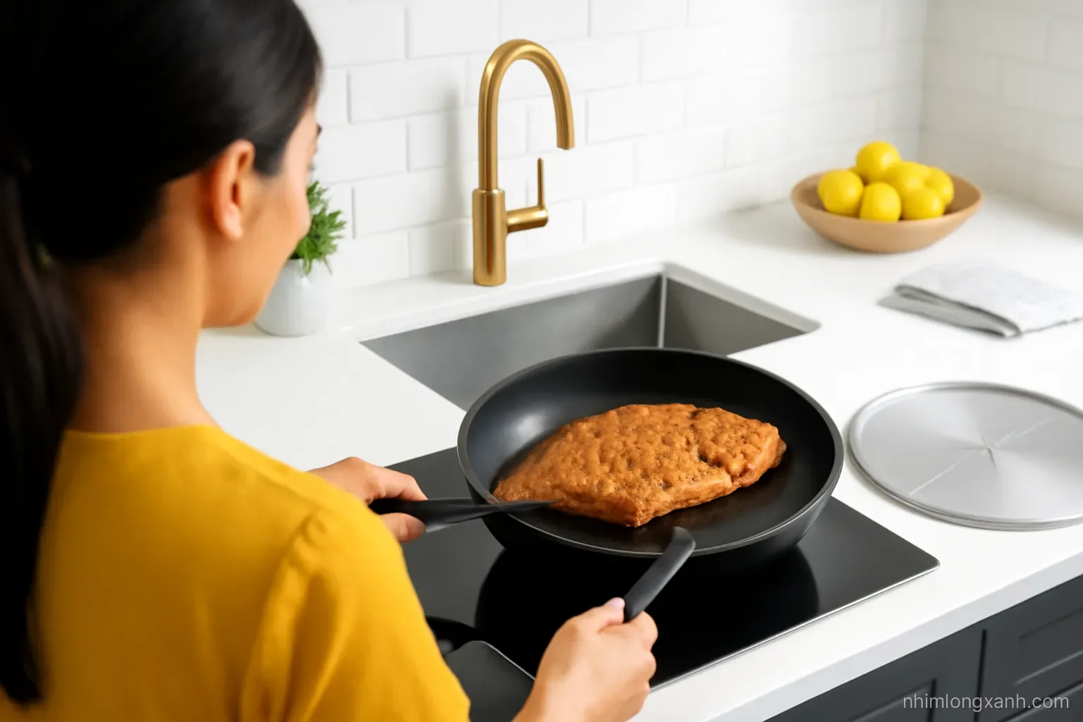 Over-the-shoulder shot of a Vietnamese woman flipping a crispy golden whole tilapia with a spatula in a black skillet, splatter guard nearby, bright modern kitchen, gold and slate tones