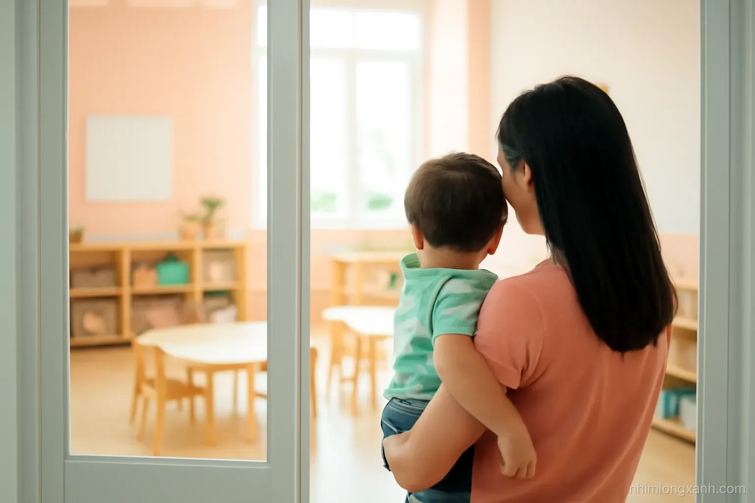 Over-the-shoulder view of a Vietnamese mother holding her toddler son, peering through a glass door into a sunlit preschool classroom with low wooden shelves, warm peach and mint tones