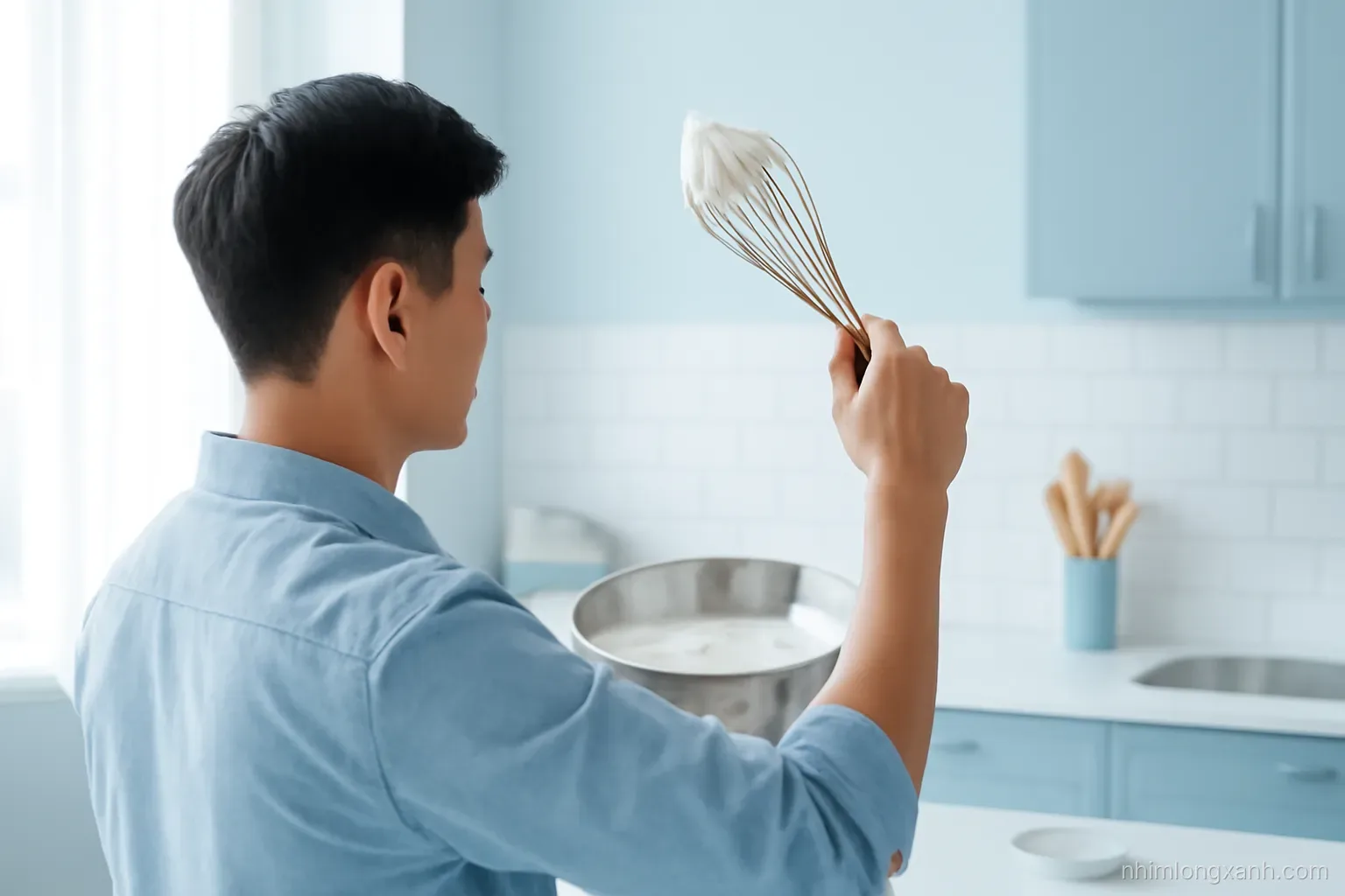 Over-the-shoulder shot of a young Vietnamese man holding up a whisk with stiff glossy white peaks above a steel bowl, bright airy studio kitchen, soft blue palette