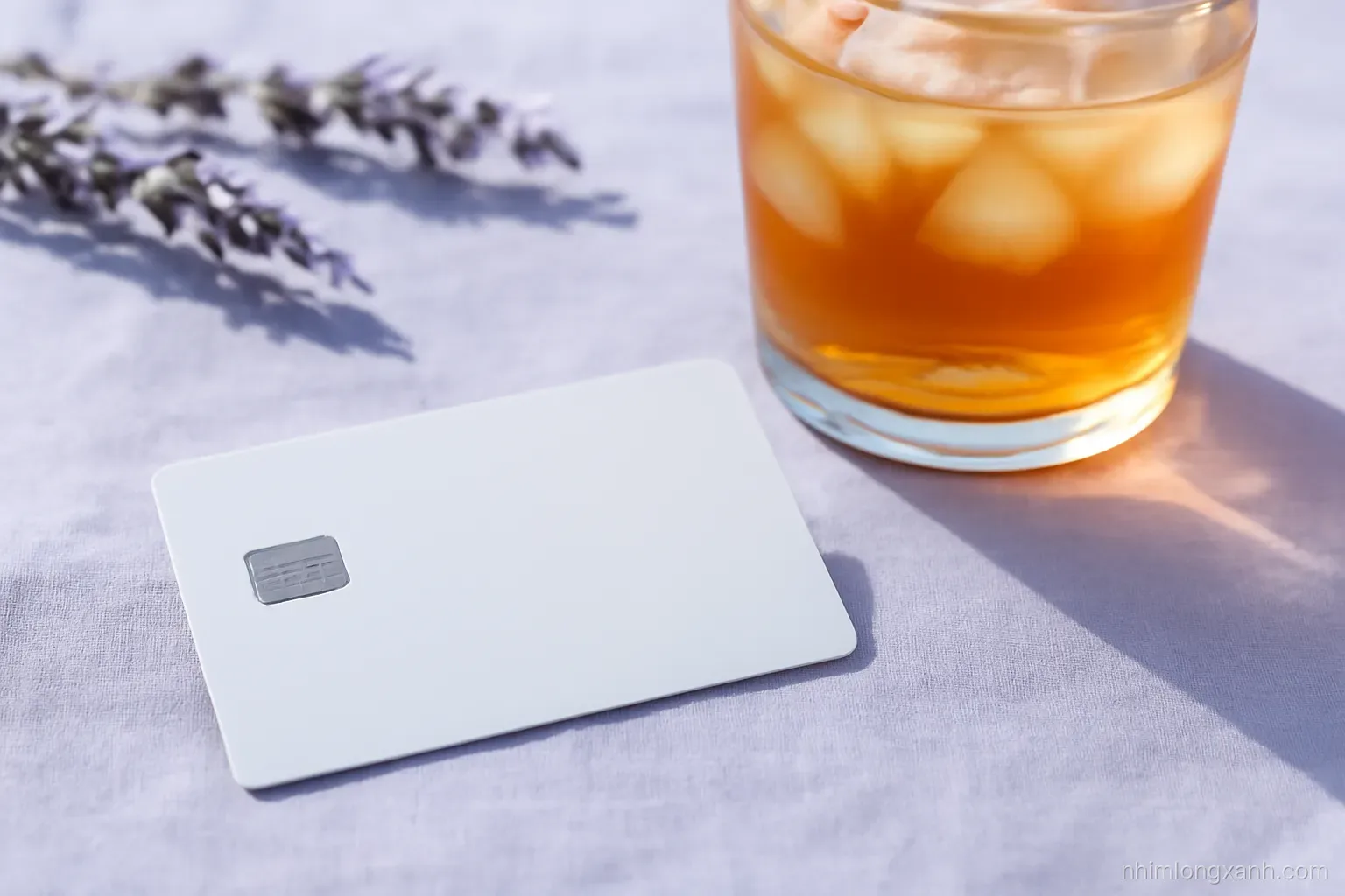 Macro close-up of a plain unbranded credit card resting on a linen tablecloth beside a cup of iced tea, crisp morning light, lavender and soft white palette