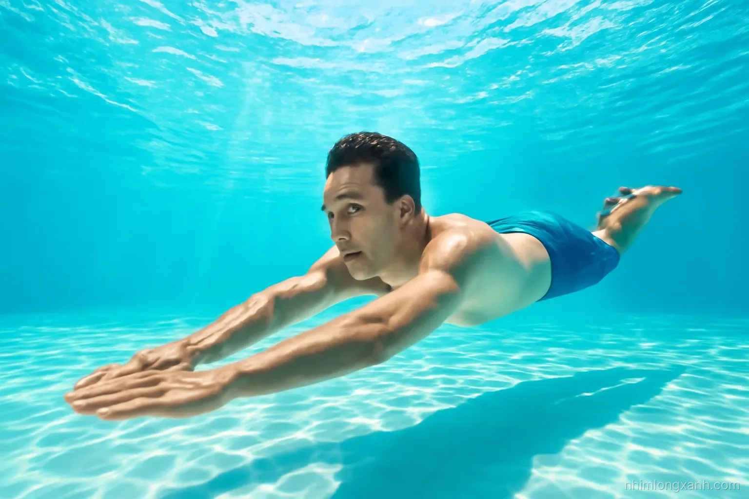 Underwater wide shot of a Vietnamese adult man gliding forward in a clear turquoise swimming pool, sunlight rays dancing through the water, bright aqua and white palette