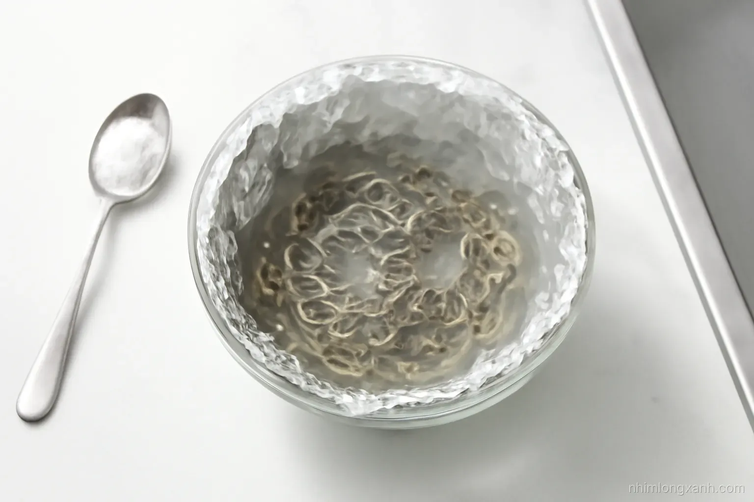 Top-down shot of a glass bowl lined with aluminium foil holding tarnished silver chains in steaming water, a spoon of white powder beside it, bright kitchen daylight, white and steel tones