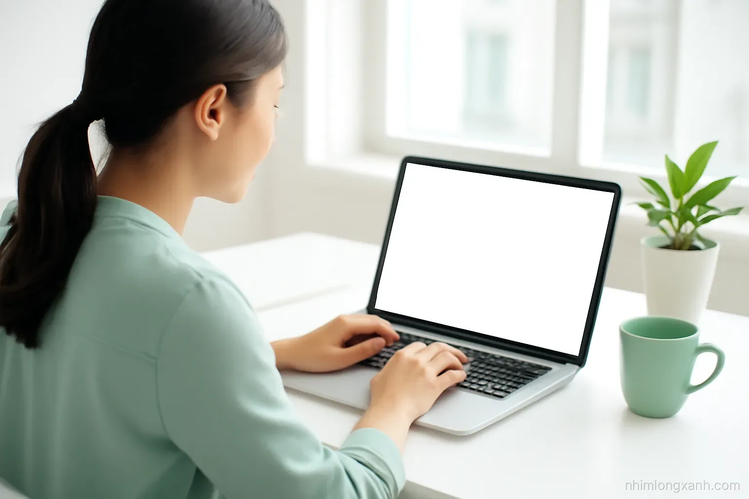 Young Vietnamese office worker typing on a laptop with a blank glowing screen at a bright window desk, over-the-shoulder shot, soft morning light, white and mint tones