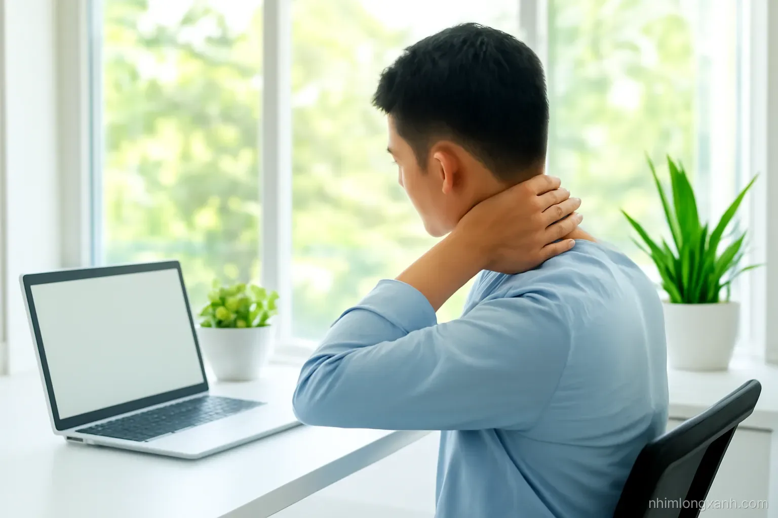 Over-the-shoulder view of a Vietnamese office worker in his thirties stretching his neck beside a desk near a large window, blank laptop, fresh green plants, bright daylight