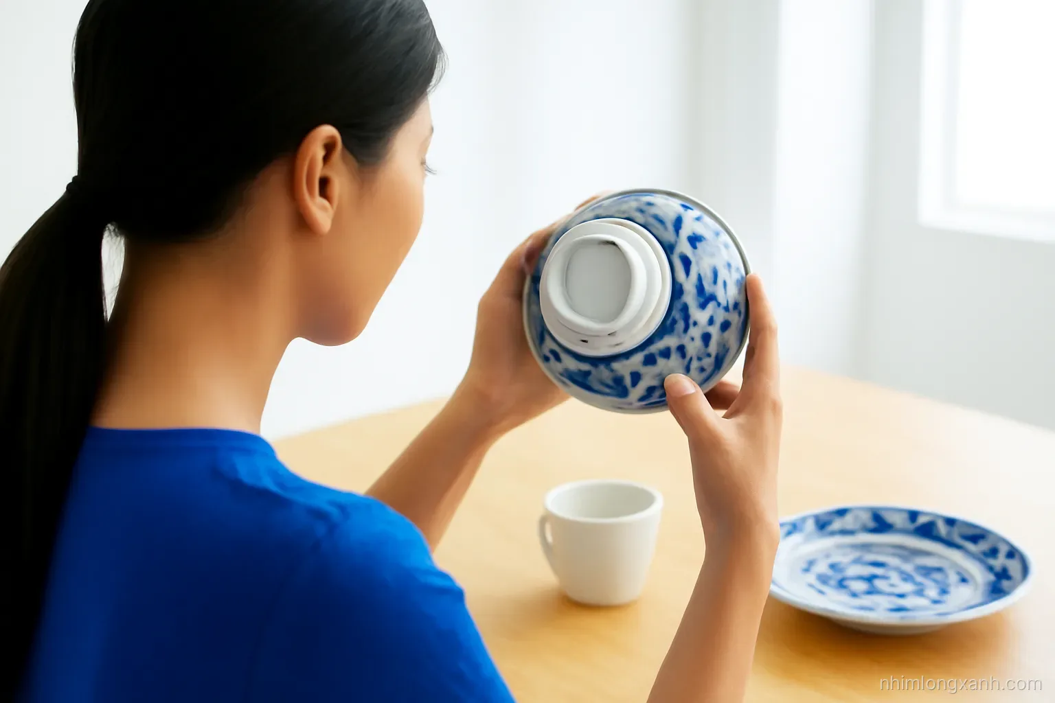 Over-the-shoulder view of a Vietnamese woman turning a blue and white porcelain bowl upside down to inspect its foot ring at a bright wooden table, cobalt blue and white palette