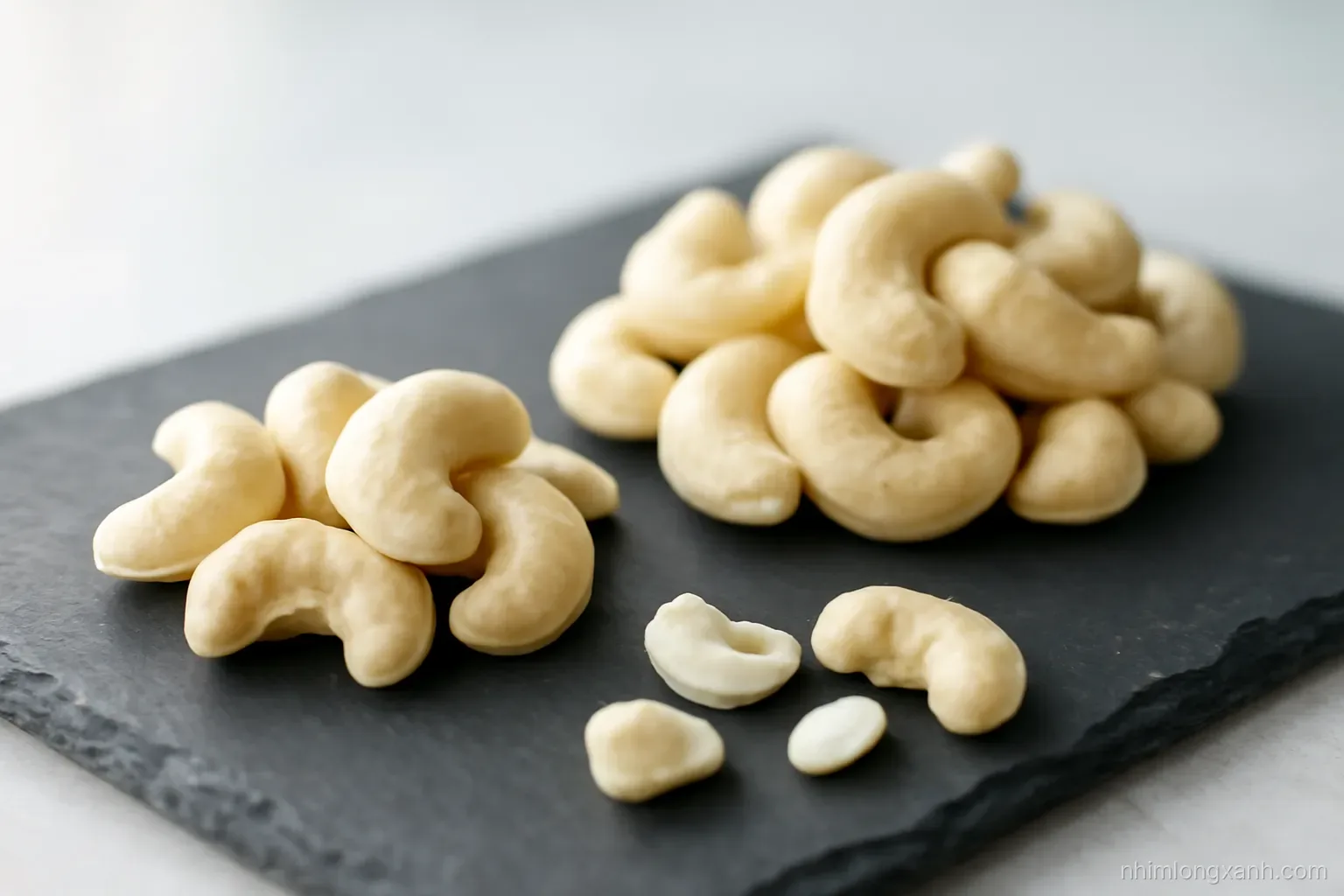 Close-up macro of whole ivory cashew kernels in two neat piles of different sizes on a slate plate, a few broken halves in front, soft side light, cream and charcoal tones