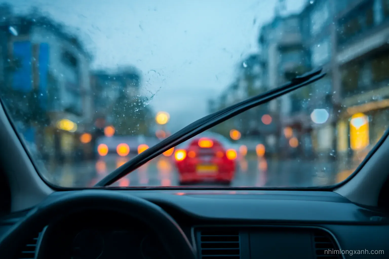 View from inside a car through a rain-streaked windshield, wipers mid-sweep, blurred red tail lights ahead on a wet Vietnamese city street at dusk, moody blue with warm bokeh