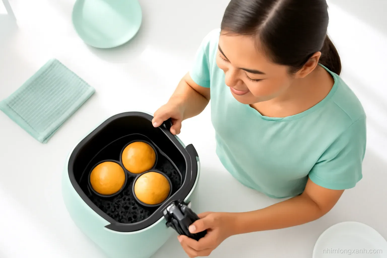 Three-quarter angle shot of a Vietnamese woman pulling out an air fryer basket holding three small golden bread rolls, bright white countertop, mint green accents, crisp morning light