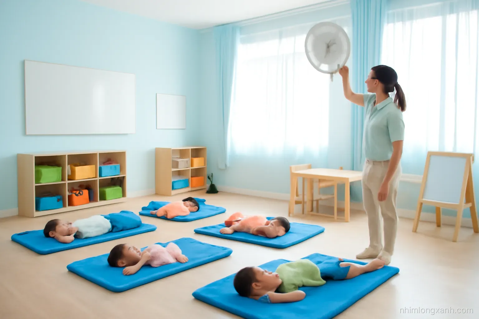 Wide angle of a bright Vietnamese kindergarten classroom at noon with small children lying on blue mats for nap time, teacher adjusting a fan, soft sunlight through curtains, pale blue tones
