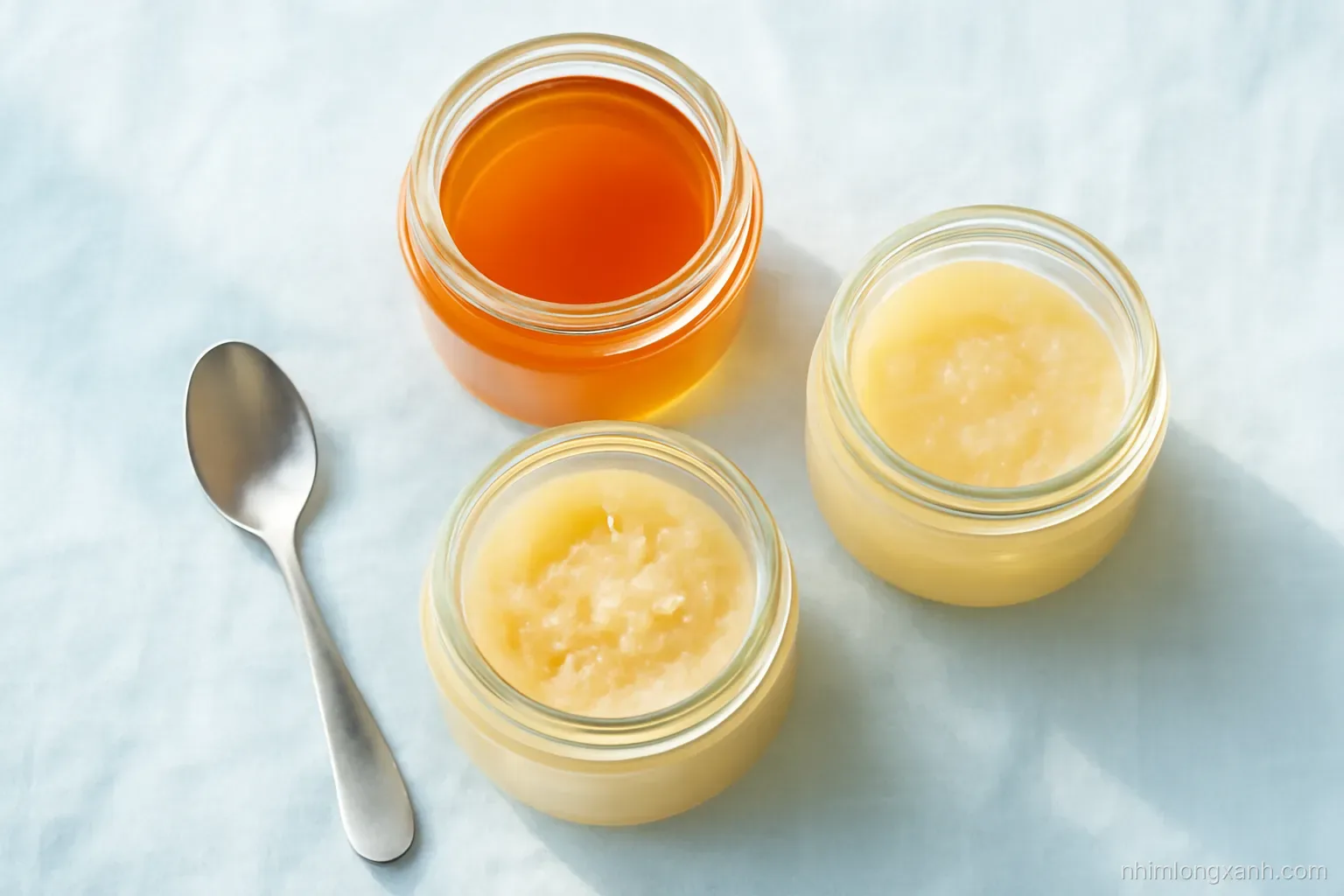 Top-down flat lay of three glass jars of honey on pale blue linen, one liquid amber, one half creamy, one fully pale crystallized, a spoon resting beside, soft morning light