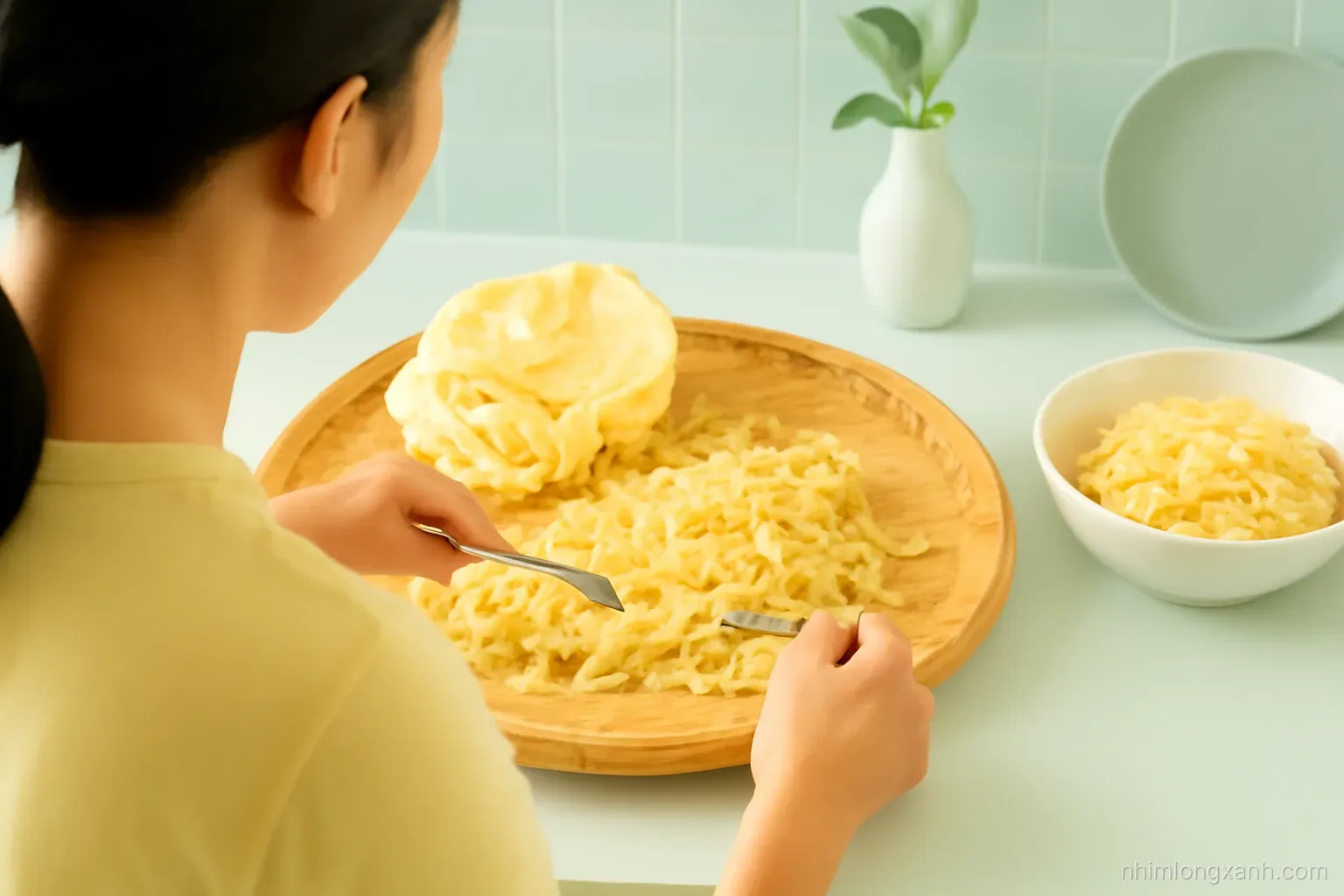 Over-the-shoulder view of a Vietnamese woman shredding boiled young jackfruit with two forks into fibrous strands on a bamboo tray, soft green kitchen light, pale yellow and leaf green palette