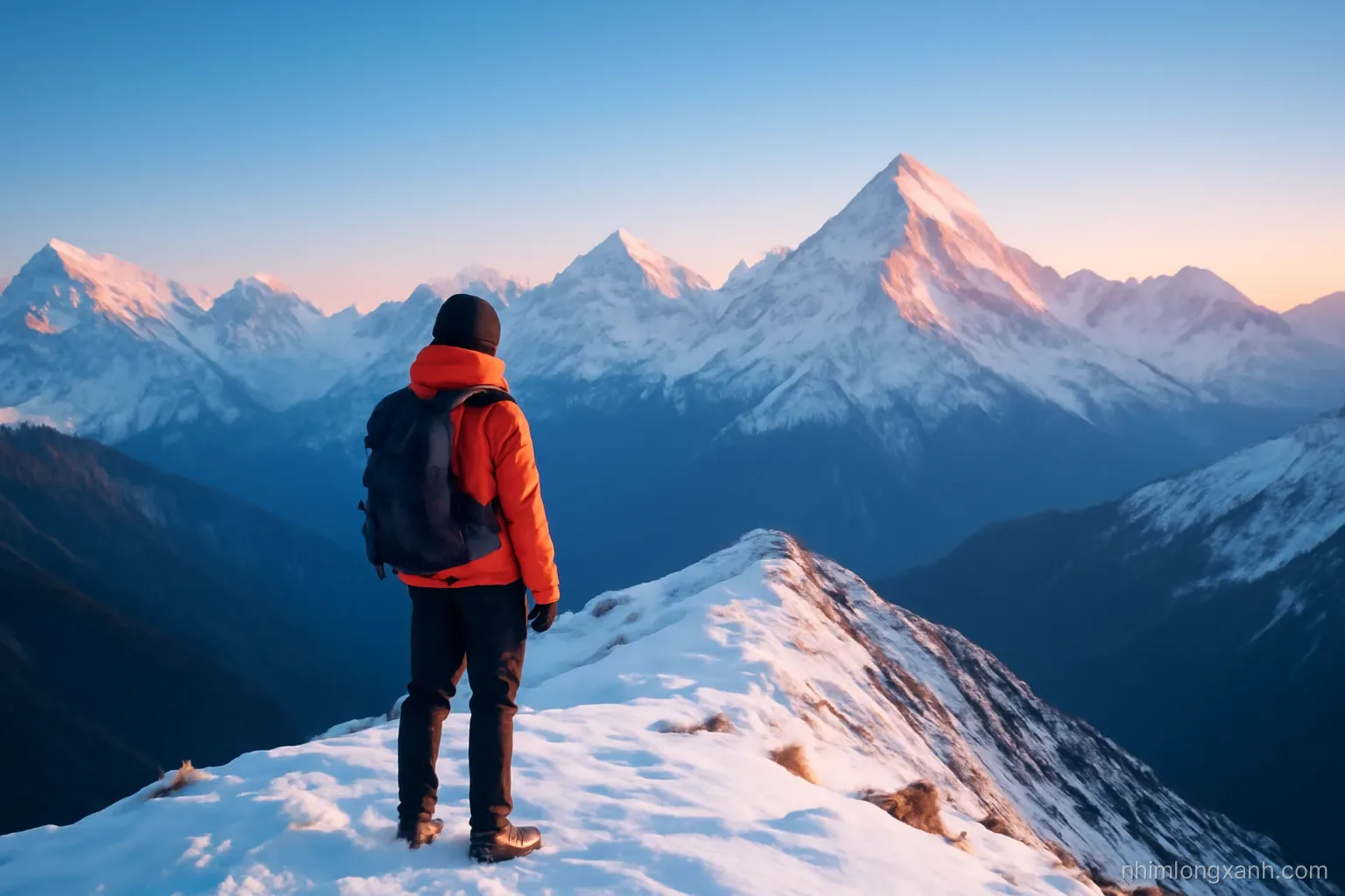 Wide shot of snow-covered Himalayan peaks at sunrise seen from a high trekking ridge, a lone Vietnamese hiker in an orange jacket in the foreground, crisp blue sky, pink alpine light
