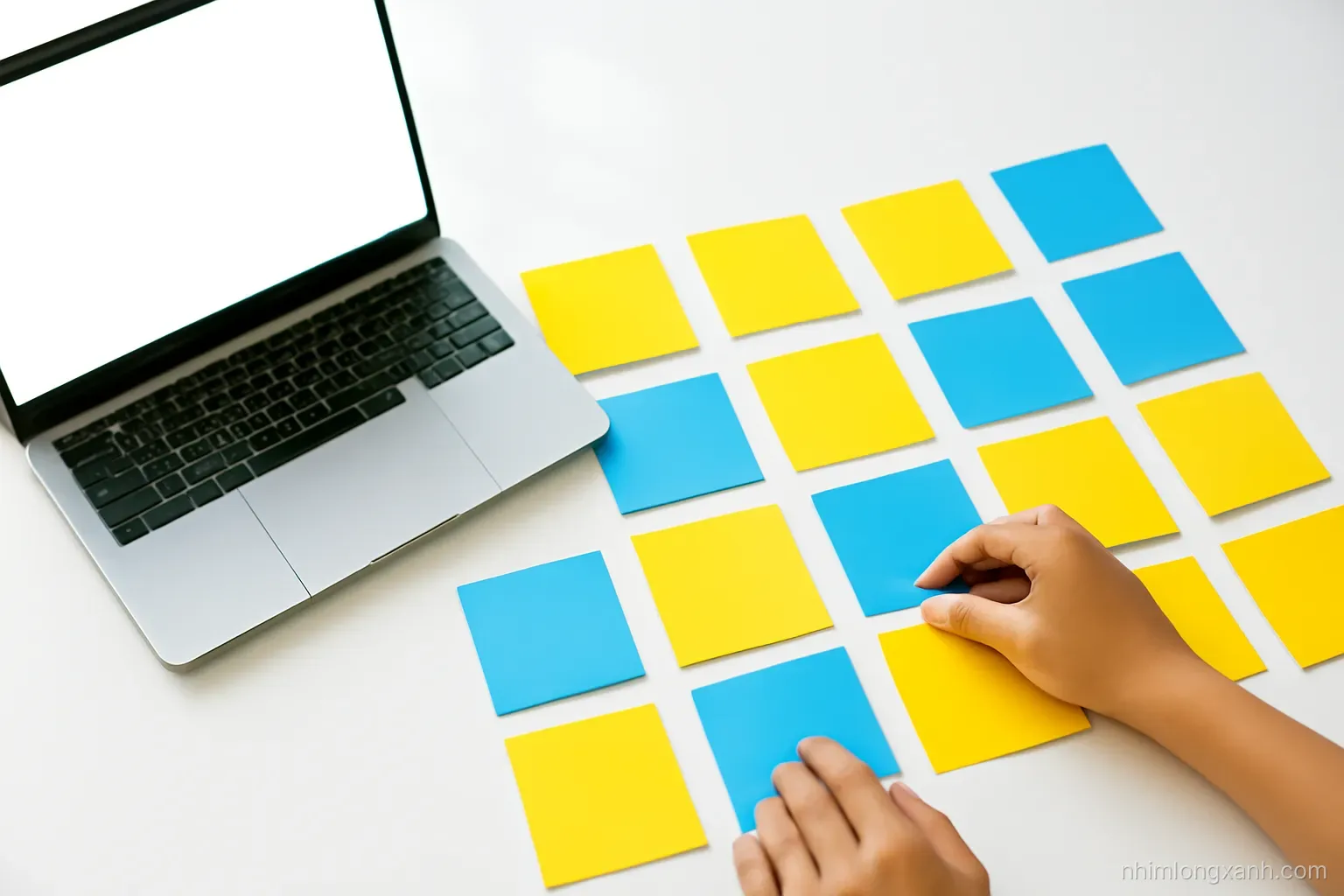 Close-up of Vietnamese hands arranging colorful paper cards into neat rows and columns on a white table beside a laptop with blank glowing screen, overhead daylight, bright yellow and blue palette