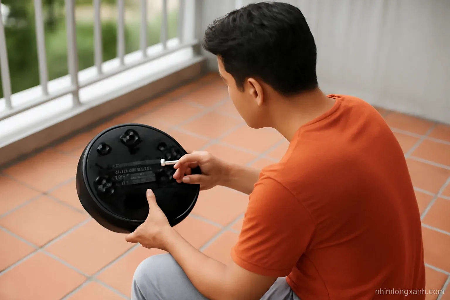 Over-the-shoulder view of a Vietnamese man kneeling on a tiled balcony, cleaning an upturned robot vacuum brush with small scissors, soft overcast light, terracotta and steel grey tones
