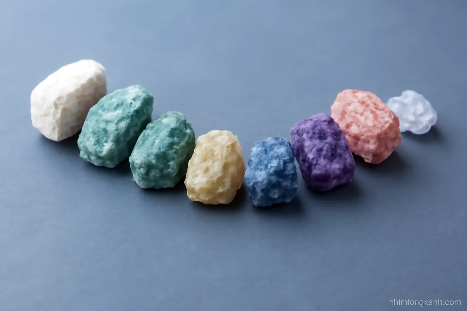 Macro close-up of ten raw mineral specimens arranged in a curved line on smooth slate, from chalky white talc to a tiny sparkling crystal, crisp side lighting, cool grey and violet tones