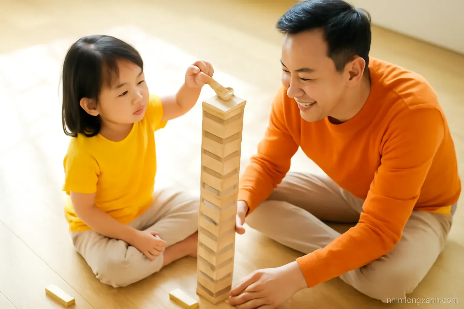 Top-down view of a Vietnamese child and father building a tall wooden block tower on a light oak floor, small hands placing a piece, bright natural light, cheerful yellow and orange tones