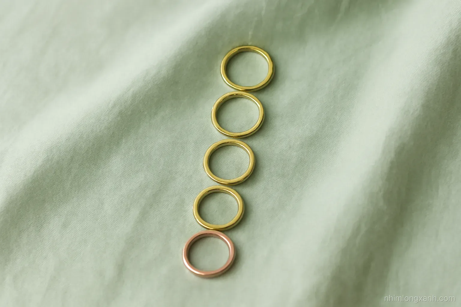 Top-down flat lay of five gold rings in graded shades from deep yellow to pale rose on a sage green linen cloth, soft diffused daylight, minimal modern styling