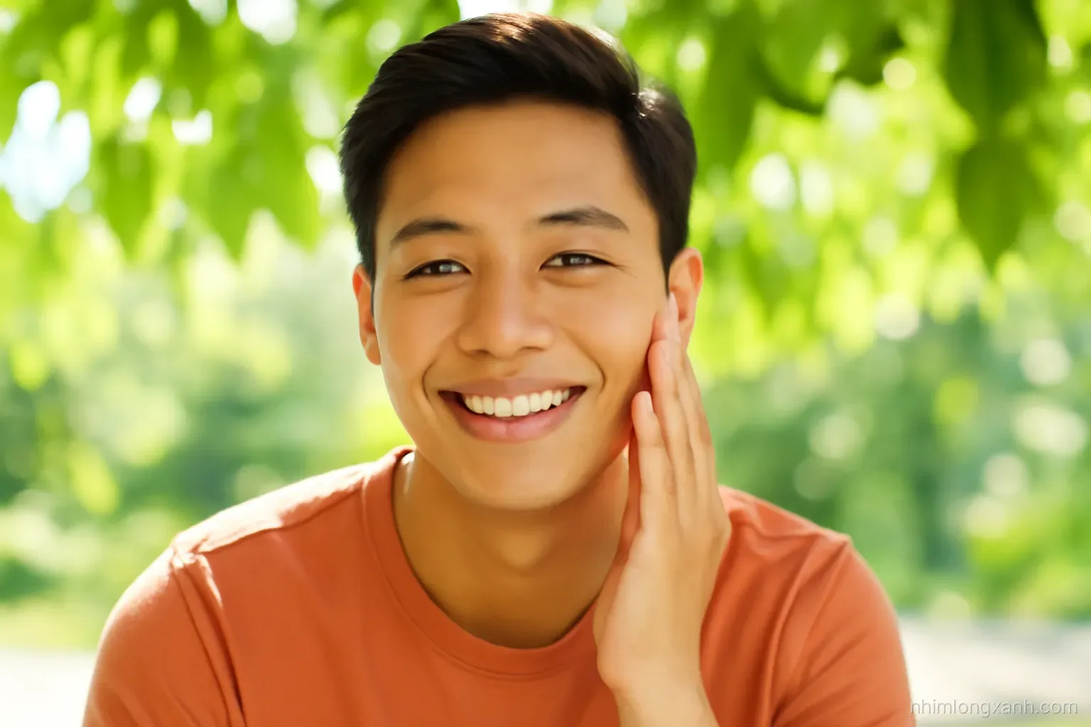 Close-up portrait of a smiling young Vietnamese man with clear natural skin touching his cheek, sitting outdoors under leafy shade, gentle backlight, vibrant fresh green and warm skin tones