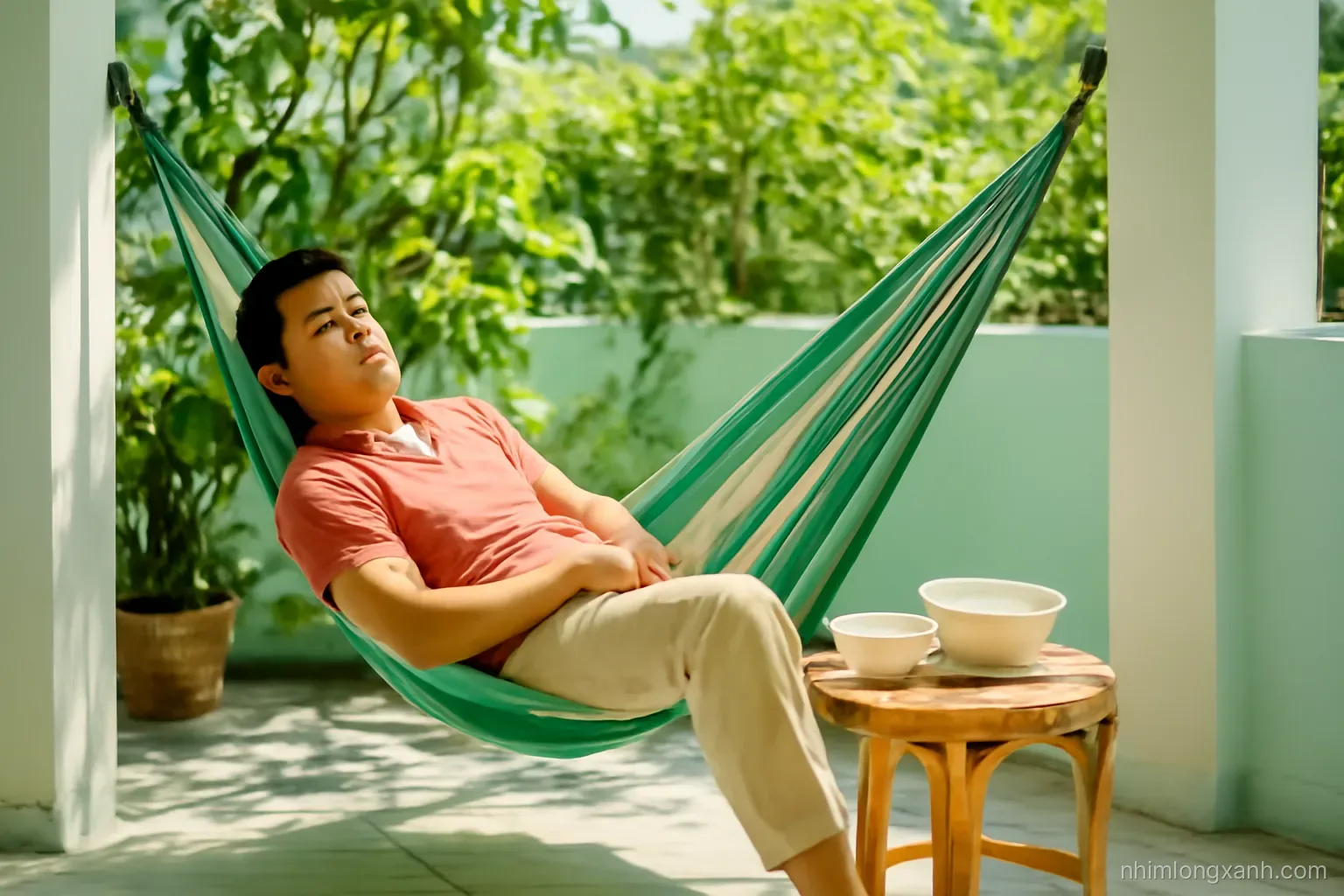 Wide shot of a Vietnamese man dozing in a hammock on a shaded garden terrace after lunch, empty bowls on a small rattan table nearby, dappled midday sunlight, mint green and coral tones