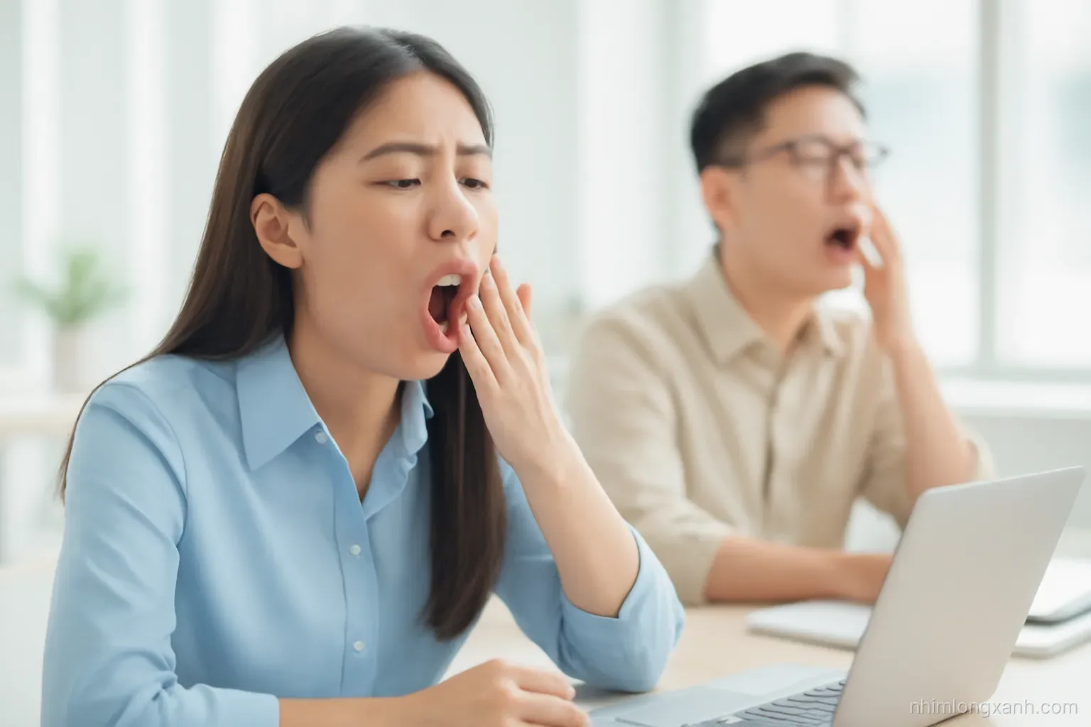 Close-up of a young Vietnamese office worker yawning widely at a sunny co-working desk while a colleague behind her also yawns, soft pastel tones, shallow depth of field