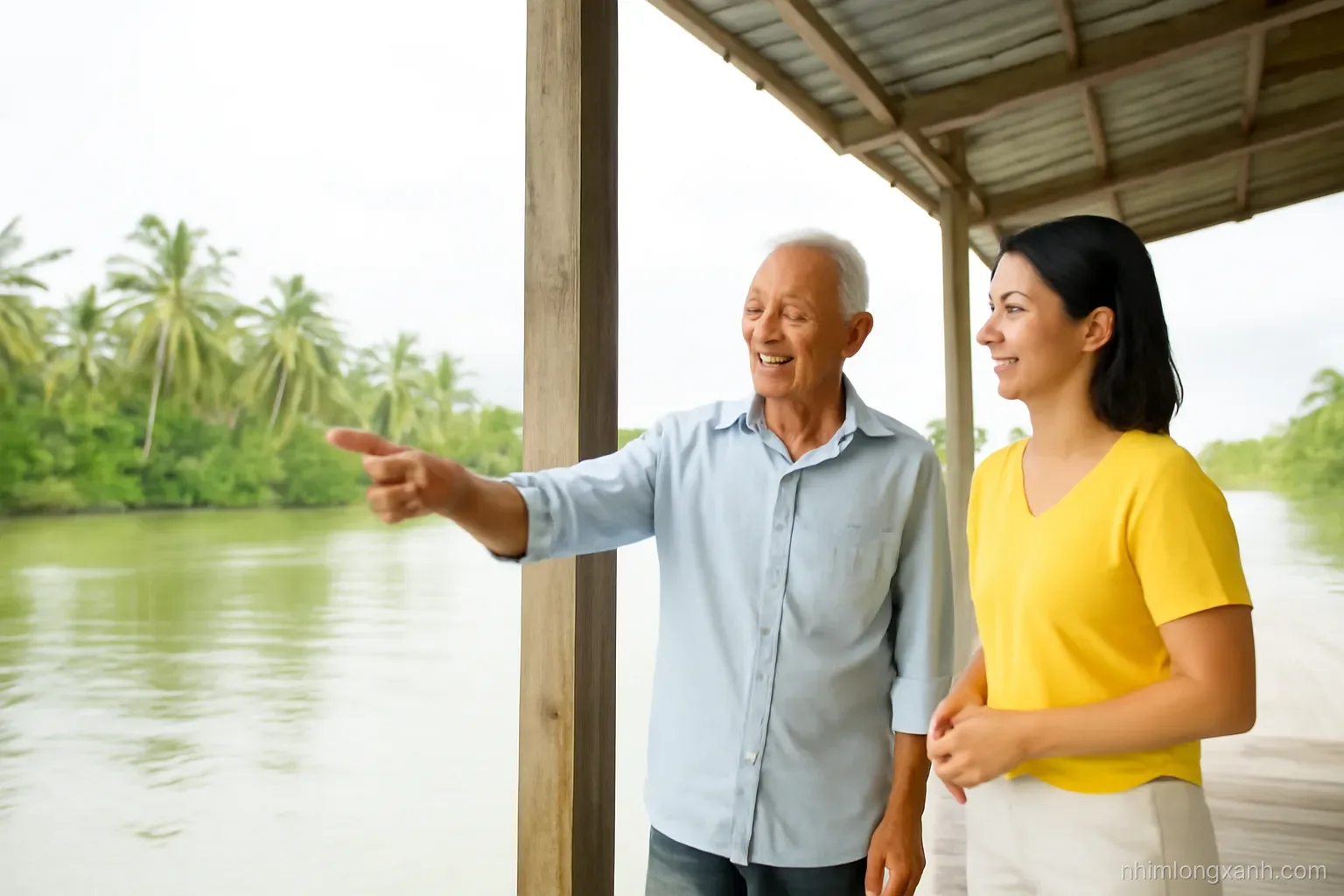 Wide shot of an elderly Vietnamese man pointing to a high water mark on a stilt house pillar while talking to a younger woman, river delta and coconut palms behind, overcast silver light